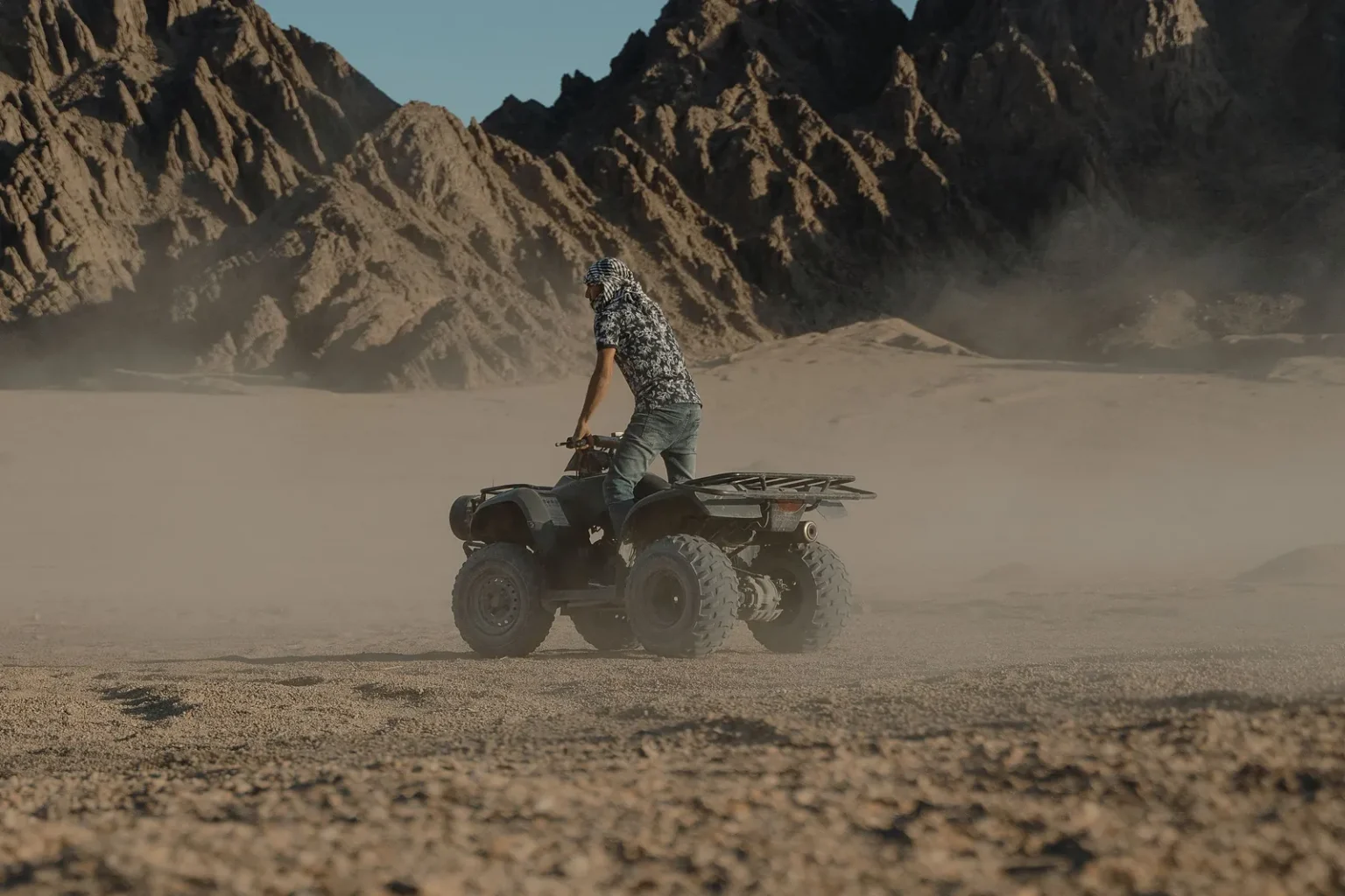Rider standing on a quad bike on a dusty desert track below rocky mountains