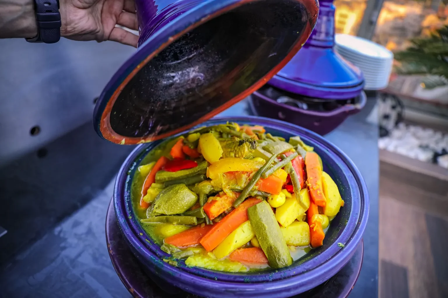 Lid lifted from a blue tagine full of cooked carrots, courgettes and potatoes