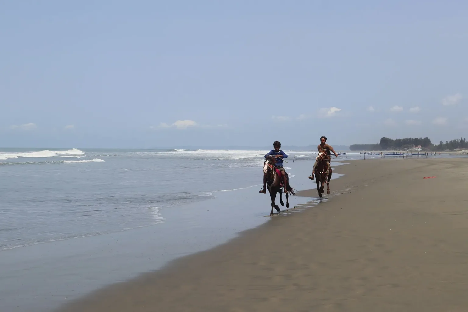 Two riders on horseback at the water's edge of a long sandy beach