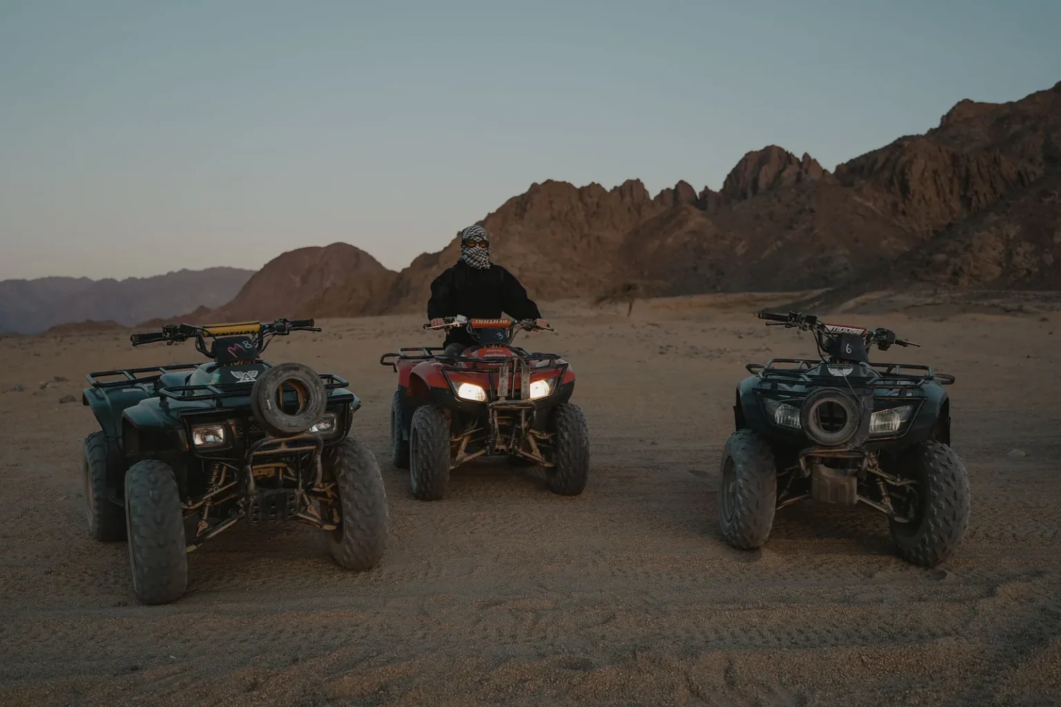 Three quad bikes parked on a desert track below rocky mountains at dusk