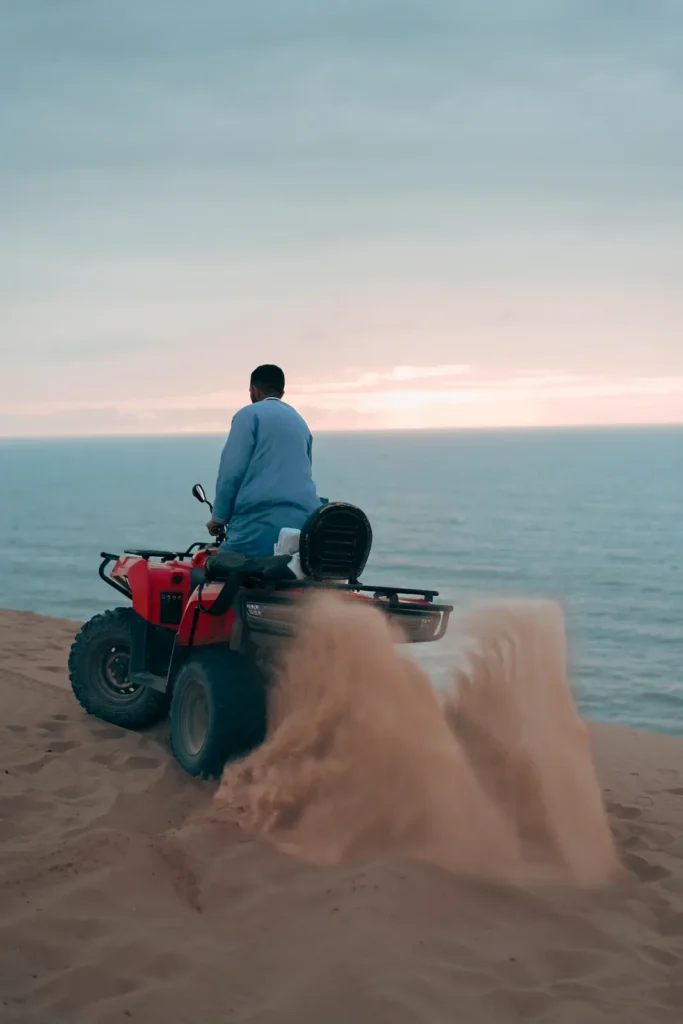 Quad rider spraying sand on a dune at sunset above the ocean